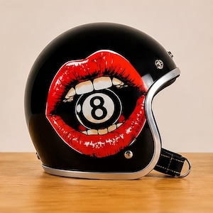 Könnte beinhalten: Ein glänzend schwarzer Jethelm für Motorräder mit einer Grafik von leuchtend roten Lippen, die in eine schwarz-weiße Acht-Ball-Billardkugel beißen. Der Helm hat eine silberne Metallkante und einen schwarzen Kinnriemen mit einem D-Ring-Verschluss.