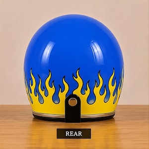 Può includere: Un casco da moto blu lucido caratterizzato da un vivace motivo a fiamme gialle lungo il bordo inferiore. Il casco include un cinturino in pelle nera con un bottone a pressione in metallo argentato sul retro e un bordo argentato alla base.