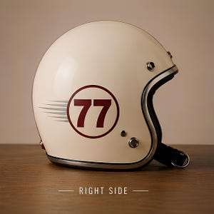 Può includere: Un casco da moto aperto color bianco sporco con una grafica circolare rosso scuro che mostra il numero 77 al centro, accompagnato da linee di velocità orizzontali grigie. Il casco presenta un bordo argentato e bottoni a pressione.