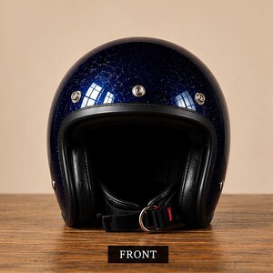 Può includere: Un casco da moto blu scuro con finitura glitterata, caratterizzato da un bordo nero attorno all'apertura, tre bottoni a pressione in metallo argentato sulla fronte e un sottogola nero con chiusura a doppio anello a D. La parola FRONT è scritta sotto.