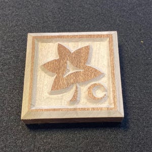 Puede incluir: Un sello de madera cuadrado con un diseño floral tallado. El diseño presenta una forma de flor central con un elemento curvo debajo. La madera tiene un color marrón claro con un diseño floral marrón más oscuro.
