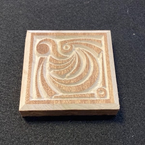 Può includere: Blocco di legno quadrato con un disegno intagliato. Il disegno presenta linee a spirale e un elemento circolare, il tutto all'interno di un bordo rettangolare. Il legno è marrone chiaro, con aree intagliate che appaiono più chiare.
