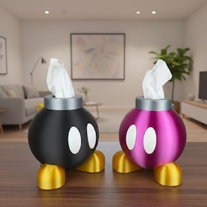 Op de afbeelding: Twee novelty tissueboxhouders in de vorm van cartoonbommen. De ene is zwart met witte ovalen en gouden voetjes, de andere is magenta met witte ovalen en gouden voetjes. Elk bevat een stapel witte tissues.