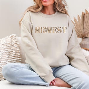 Puede incluir: Una sudadera beige de cuello redondo con la palabra MIDWEST impresa en el pecho en un estampado de leopardo marrón y tostado. La prenda tiene mangas largas y cuello, puños y dobladillo acanalados, ofreciendo un ajuste estándar y cómodo.