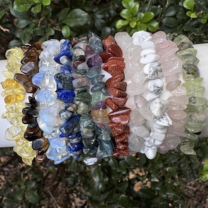 May include: A collection of gemstone chip bracelets in various colors, including yellow, brown, blue, purple, red, white, pink, and green. The bracelets are displayed on a white cylindrical surface against a backdrop of green foliage.