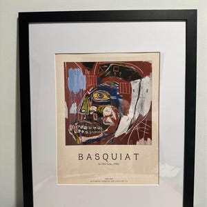 May include: Framed art print of a Basquiat abstract portrait. The artwork uses bold brushstrokes and a mix of colours, including red, blue, and yellow. The print is set in a black frame with a white mount.