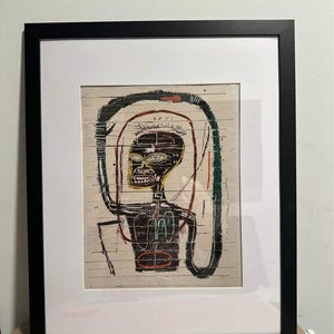 May include: Framed artwork depicting a stylized figure with a black head and torso, yellow face, and red and green accents. The artwork is set against a light-coloured, textured background and framed in a black frame with a white mount.