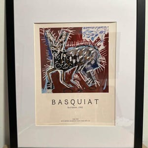 May include: Framed art print of Basquiat's "Red Rabbit, 1982." The artwork depicts a rabbit in black, white, and blue against a maroon backdrop. The print is presented with a white mount and a black frame, suitable for wall display.