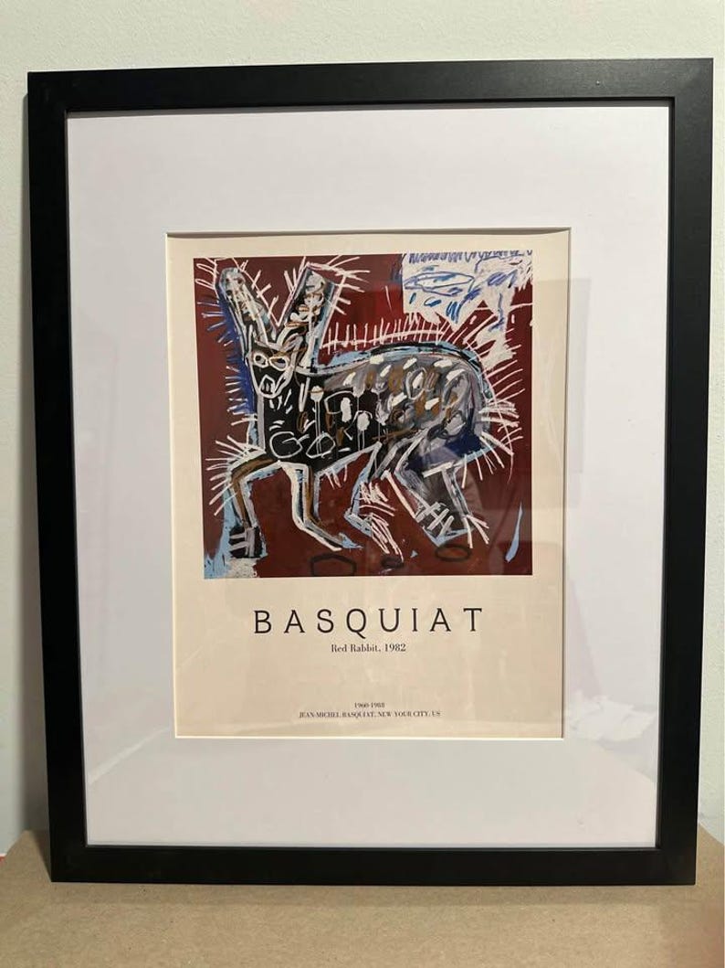 May include: Framed art print of Basquiat's "Red Rabbit, 1982." The artwork depicts a rabbit in black, white, and blue against a maroon backdrop. The print is presented with a white mount and a black frame, suitable for wall display.