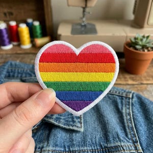 May include: A heart-shaped embroidered patch with a rainbow design. The patch features a white border and is held over a denim jacket. The rainbow colors include red, orange, yellow, green, blue, and purple.