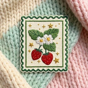 May include: A rectangular embroidered patch with a scalloped dark green border on a cream background. The design features two red strawberries with green leaves, two white flowers with yellow centers, and several small gold embroidered stars.