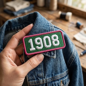 May include: A rectangular embroidered patch with the year "1908" in white, set against a green background and a pink border. The patch is displayed on a blue denim jacket, showcasing a vintage aesthetic.