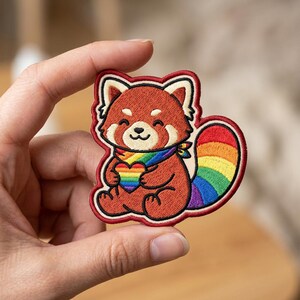 May include: An embroidered patch featuring a red panda with rust-colored fur, cream-colored facial markings, and a black nose. The red panda wears a rainbow-colored scarf, holds a rainbow heart, and has a large, striped rainbow-colored tail.
