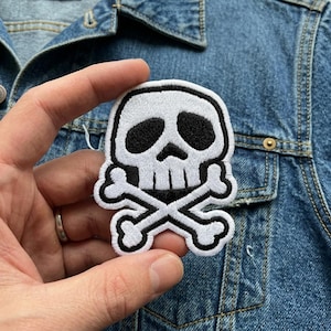 May include: Embroidered patch featuring a white skull with black eye sockets and a black outline. The skull is positioned above two crossed white bones, all with a black outline. The patch is held against a blue denim jacket.