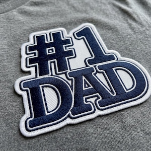May include: Close-up of a gray t-shirt with a navy blue and white embroidered patch. The patch reads "#1 DAD" in a bold, stylized font, with a white border. The fabric of the shirt has a textured appearance.