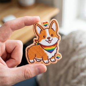 May include: An embroidered patch featuring a brown and white Corgi dog wearing a rainbow-striped bandana. The design includes a small rainbow heart above the dog's head and a rainbow-colored paw print near the bottom right, all outlined in tan thread.
