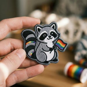 May include: An embroidered iron-on patch featuring a gray and white raccoon with black eye markings. The raccoon has a striped tail with a rainbow accent and is holding a small rainbow flag. The patch has a dark gray border and detailed stitching.
