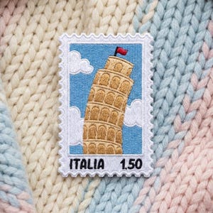 May include: A rectangular embroidered patch shaped like a postage stamp featuring the Leaning Tower of Pisa in tan thread against a light blue sky with white clouds and a small red flag. The text ITALIA 1.50 is stitched in black at the bottom.