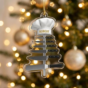 May include: A clear acrylic Christmas tree-shaped ornament featuring a white chef hat at the top with the name ROBERT printed in gold. Below the hat, a vertical strip holds five various kitchen knives and a spatula, all arranged in a tree shape.