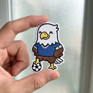 May include: An embroidered patch featuring a cartoon bald eagle wearing a blue soccer jersey with a white captain's armband marked with the letter C. The eagle has brown wings and body, a yellow beak, and stands with one foot on a soccer ball.