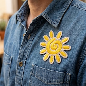 May include: A denim shirt with a yellow embroidered sun patch on the chest. The sun design features a spiral center and petal-like rays, outlined in a cream color. The shirt has a button-down front and a chest pocket.