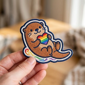 May include: An embroidered patch featuring a brown otter with a smiling face, holding a rainbow-colored heart. The otter is positioned above a stylized rainbow swirl design, all outlined in a dark blue border against a white background.