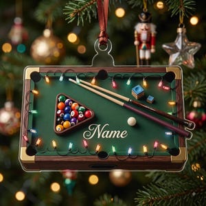 May include: A rectangular Christmas tree ornament shaped like a pool table with a green felt surface, wooden rails, and gold corner accents. It features a rack of colorful billiard balls, two cues, a white cue ball, chalk, and the word Name.