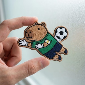 May include: An embroidered patch featuring a brown capybara dressed as a soccer goalkeeper. The capybara wears a green jersey with a white captain's armband marked with a yellow C, white gloves, and dark blue shorts, diving toward a soccer ball.