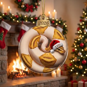 May include: A round acrylic Christmas ornament featuring a coiled white and yellow snake wearing a small red and white Santa hat with the name LEO printed on the brim. The ornament has a gold hanging cord and the year 2026 engraved at the top.