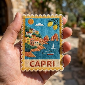 May include: A rectangular embroidered patch shaped like a postage stamp with a scalloped border. The design features a coastal landscape with colorful buildings, a blue sea, sailboats, a yellow sun, and lemons. The word CAPRI is stitched in red.
