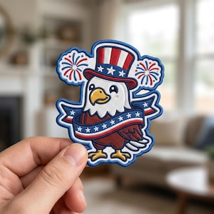 May include: An embroidered patch featuring a cartoon bald eagle wearing a red, white, and blue striped top hat. The eagle is surrounded by two stylized fireworks and is wrapped in a banner decorated with white stars on a blue background.