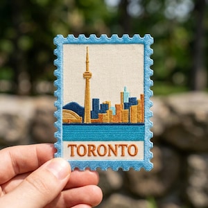 May include: A rectangular embroidered patch shaped like a postage stamp with a light blue border. The design features the Toronto skyline, including the CN Tower in gold, against a cream background, with the word TORONTO in orange text below.