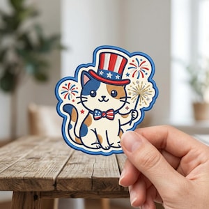 May include: An embroidered patch featuring a calico cat wearing a red, white, and blue top hat with stars and a matching bow tie. The cat holds a gold sparkler, surrounded by red and blue firework illustrations on a cream-colored background.
