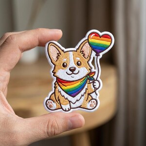 May include: An embroidered patch featuring a tan and white Corgi dog sitting down. The dog wears a rainbow-colored bandana around its neck and holds a string attached to a heart-shaped balloon with rainbow stripes. The patch has a white border.