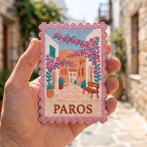 May include: A rectangular embroidered patch shaped like a postage stamp with a pink border. The design features a Mediterranean street scene in Paros with tan stone buildings, a wooden bench, and vibrant pink bougainvillea flowers. The word PAROS is embroidered in brown at the bottom.