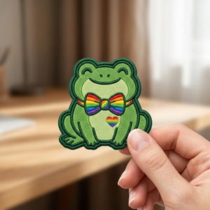 May include: An embroidered patch shaped like a green frog with a smiling face. The frog wears a rainbow-striped bow tie and has a small rainbow heart embroidered on its chest. The patch features dark green outlines and a light green belly.