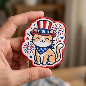May include: An embroidered patch featuring a light orange cat wearing a red, white, and blue striped top hat with stars and a matching blue bandana with white stars. The design includes red and blue firework bursts on a white background.
