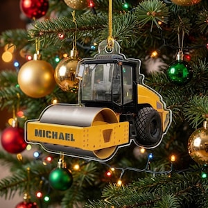 May include: A flat, acrylic Christmas tree ornament shaped like a yellow and black road roller construction vehicle. The front drum of the roller features the name MICHAEL printed in bold black capital letters. It hangs from a gold string.