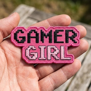 May include: An embroidered patch with a pink border featuring the text GAMER GIRL in a black pixelated font. The word GAMER is on the top line and GIRL is on the bottom line, both rendered in a blocky, digital-style typeface with pink stitching.