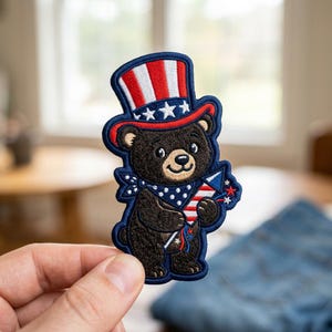 May include: An embroidered patch featuring a brown bear wearing a red, white, and blue striped top hat with white stars. The bear has a blue bandana with white polka dots around its neck and is holding a red, white, and blue firework rocket.