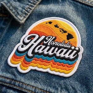 May include: An embroidered patch featuring a sunset design with yellow, orange, and blue waves, topped with a yellow sun containing black island silhouettes. The text Honolulu Hawaii is stitched in white with a black outline over the design.