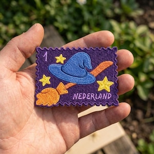 May include: A rectangular embroidered patch with a purple background and scalloped edges. It features a blue witch hat, an orange broomstick, three yellow stars, the number 1, and the text NEDERLAND stitched in light purple thread.