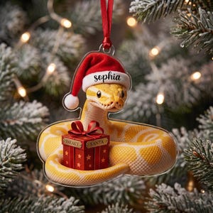 May include: A clear acrylic Christmas ornament featuring a yellow snake wearing a red and white Santa hat with the name Sophia printed on the brim. The snake is coiled around a red gift box with a bow, labeled with the text Sneaky Santa.
