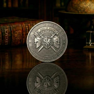 May include: A silver-colored coin with the text "Through Ages of Valor" and "Ancient Covenants." The coin features a shield with a bird and crossed swords, axes, and helmets. The coin is displayed on a reflective surface.
