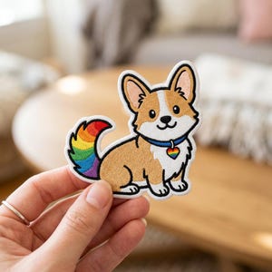 May include: An embroidered patch featuring a tan and white Corgi dog with a rainbow-colored tail and a blue collar with a rainbow heart-shaped tag. The design has a thick black outline and a white border, showcasing a cartoon-style illustration.