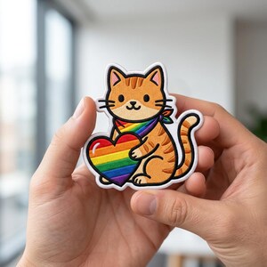 May include: An embroidered patch featuring an orange tabby cat with a rainbow-colored bandana around its neck. The cat is holding a large, heart-shaped object with horizontal stripes in the colors of the rainbow, outlined in a thick white border.