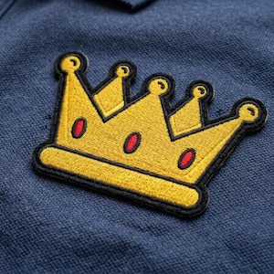 May include: Embroidered gold crown patch with red accents and a black border, affixed to a navy blue fabric. The crown design features a classic royal shape, suitable for apparel or accessories.