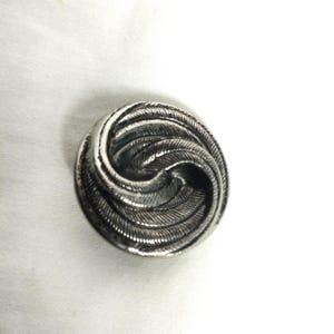May include: A circular silver-toned button featuring a raised, swirling pattern of textured lines that radiate from the center toward the outer edge, creating a dynamic, spiraling effect across the entire surface of the metallic fastener.