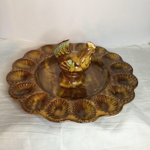 May include: A round, brown glazed ceramic egg serving platter featuring a central handle shaped like a rooster. The platter has a scalloped edge and twelve circular indentations arranged around the perimeter for holding individual eggs.