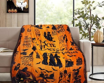 May include: Orange Halloween-themed throw blanket with black silhouettes of witches, pumpkins, bats, and text. The blanket is draped over a beige sofa. A framed Halloween picture hangs on the wall.